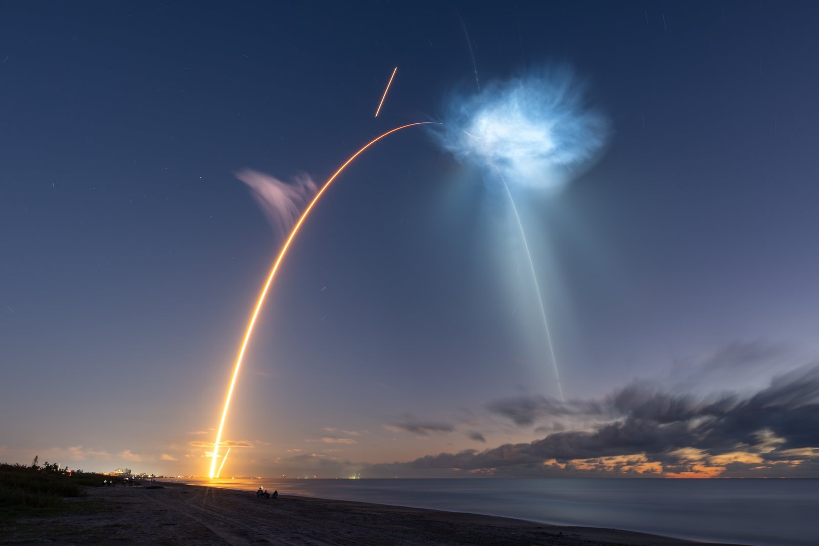 A long-exposure photo of a bright orange rocket trail curving up from the coast over the ocean at dawn, with a glowing blue plume high in the sky