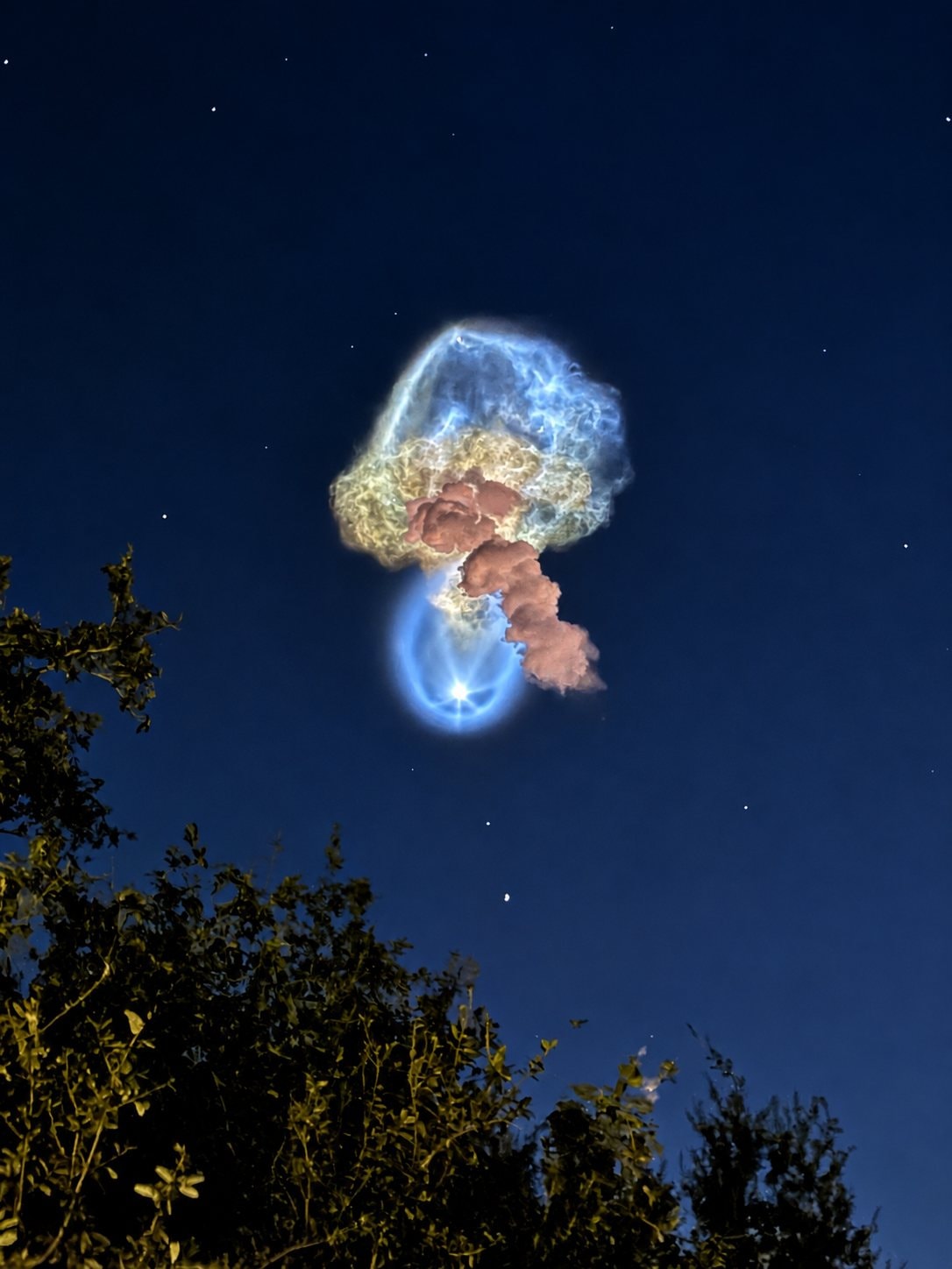 A glowing, jellyfish-shaped rocket plume in the predawn sky over Florida, with a small round blue glow at its base that looks like a stone with a star inside